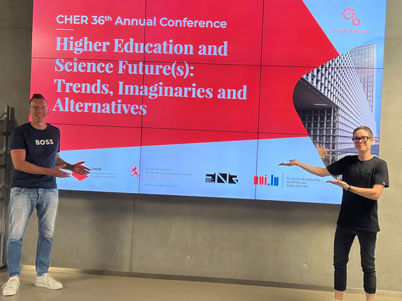 Uwe Wilkesmann (left) and Sabine Lauer (right) standing in front of the CHER conference poster.