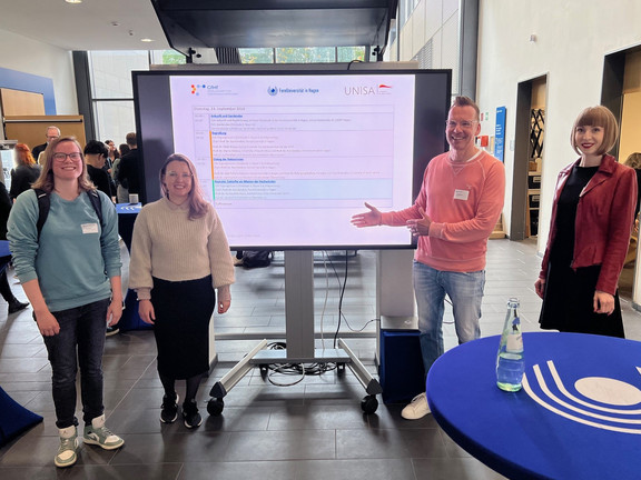 from left to right: Alina Breitzke, Olga Wagner, Uwe Wilkesmann, Viktoria Jäger in front of the screen with the programme announcement.